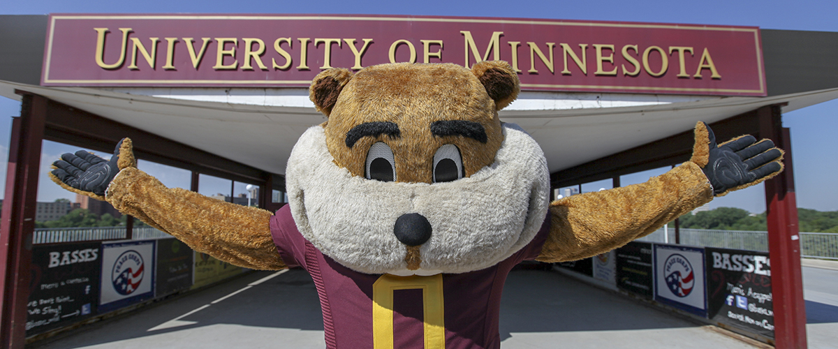 Goldy the Gopher standing with arms outstretched in front of UMN pedestrian bridge
