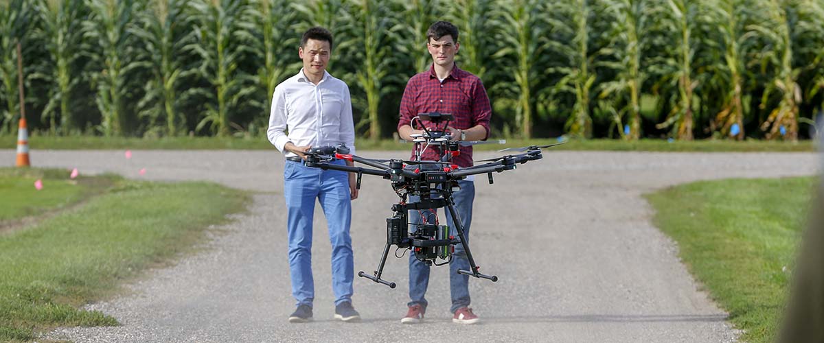 Two students stand side by side, with one operating a drone used for agricultural research.