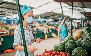 A woman with a head scarf and a blue face mask looks at watermelons and peaches at a farmers market.