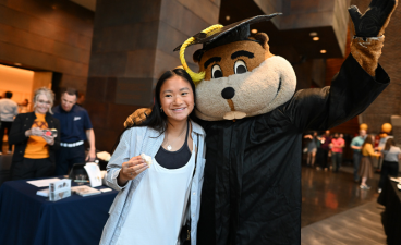 Goldy Gopher dressed in a cap and down posing for a photo with a graduating senior. 